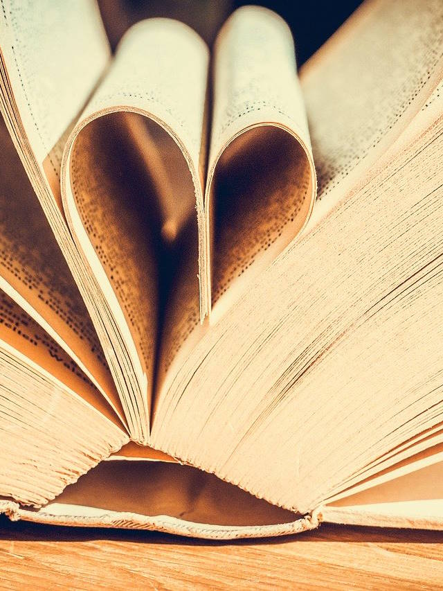 Otwartą książkę, której kartki są ułożone w kształt serca. Zdjęcie przedstawia zbliżenie na otwartą książkę, z kartkami zgiętymi tak, że tworzą kształt serca. Widoczny jest drewniany blat, na którym leży książka.