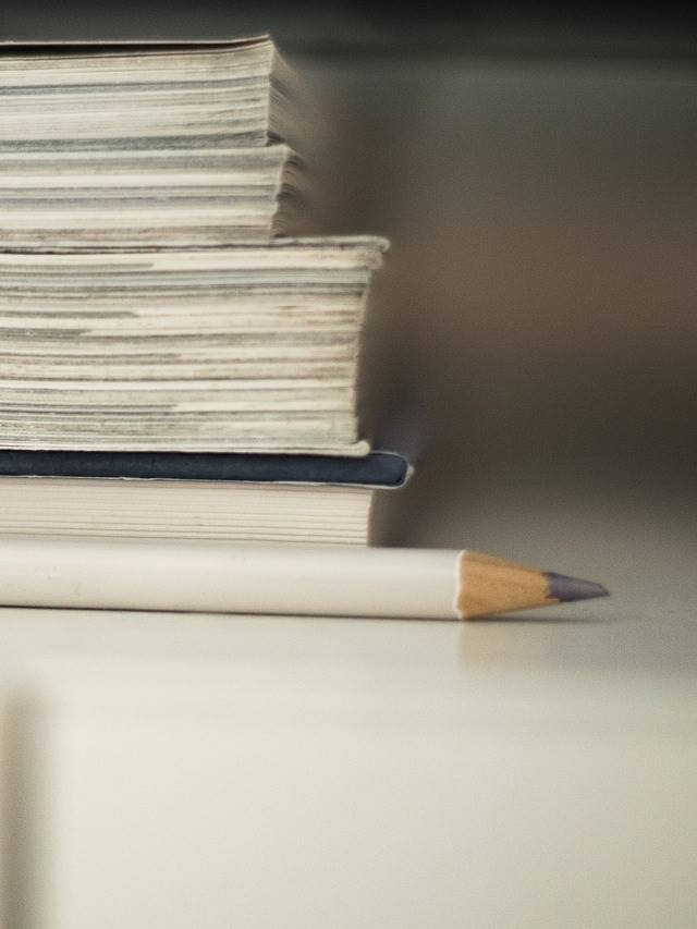 Stos magazynów i książek na biurku z leżącym obok ołówkiem. Zdjęcie przedstawia zbliżenie na stos papierowych magazynów i książek ułożonych jeden na drugim na jasnej powierzchni biurka. Obok stosu leży drewniany ołówek z niebieską końcówką.